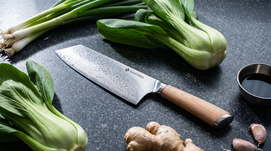 A tall Damascus-clad vegetable cleaver on a charcoal granite worktop with pak choi, spring onions and ginger
